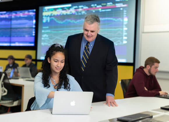 A professor and student talk in the financial technology center with stock tickers on the screens around them